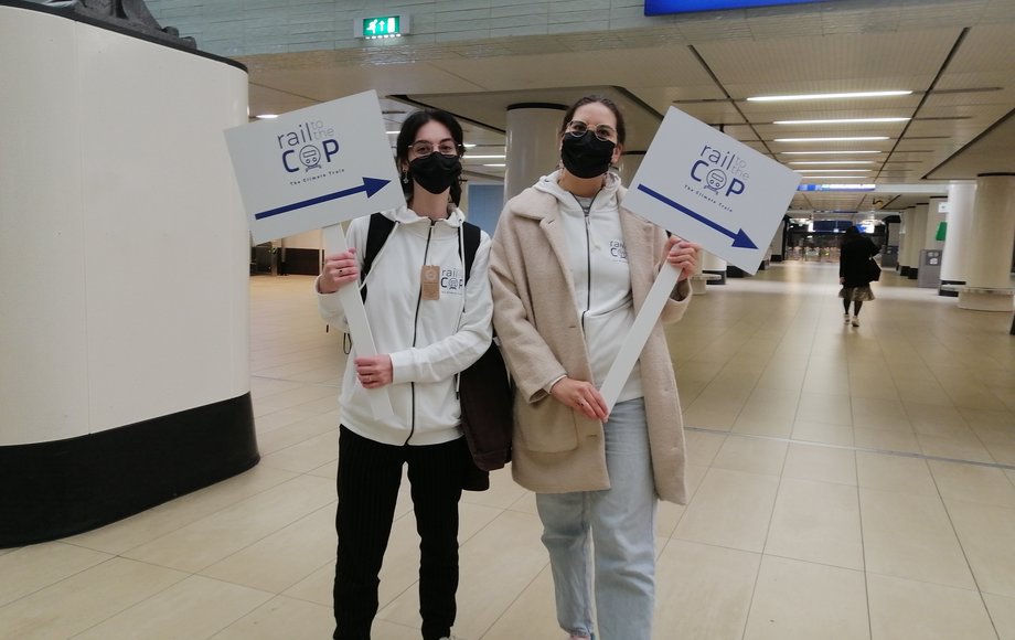 Zwei junge Mädchen halten Wegweiserschilder am Bahnhof mit Aufschrift „Rail to the COP“