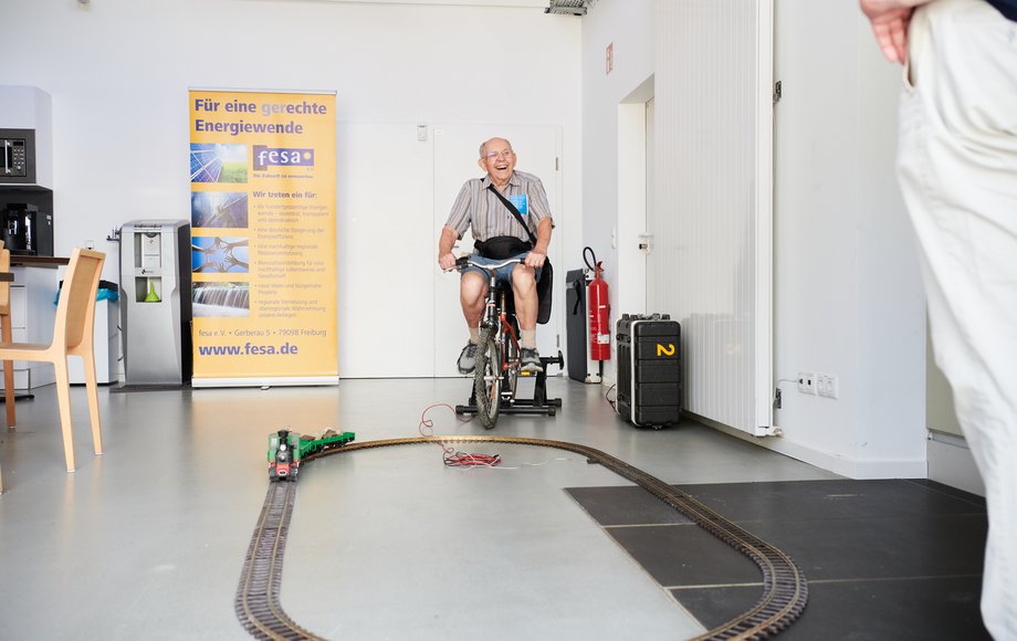 Ein älterer Mann sitzt auf einem Hometrainer, vor ihm eine Modelleisenbahn. Er tritt lachend in die Pedale 