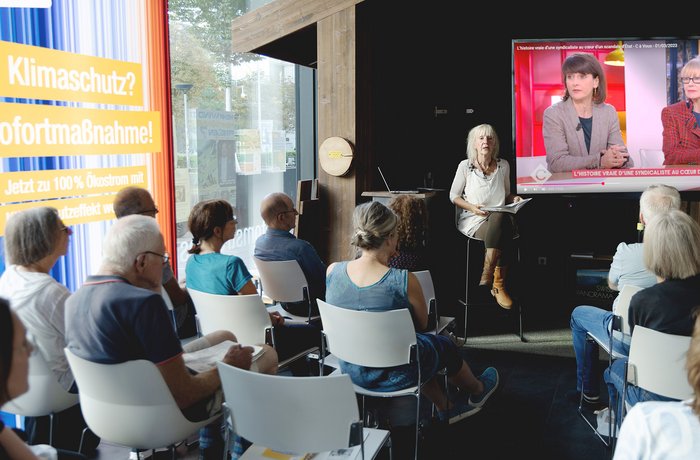 Szenerie im Freiburger Store mit Leserin Eva Stegen vor Publikum, daneben ein Monitor mit Grußwort