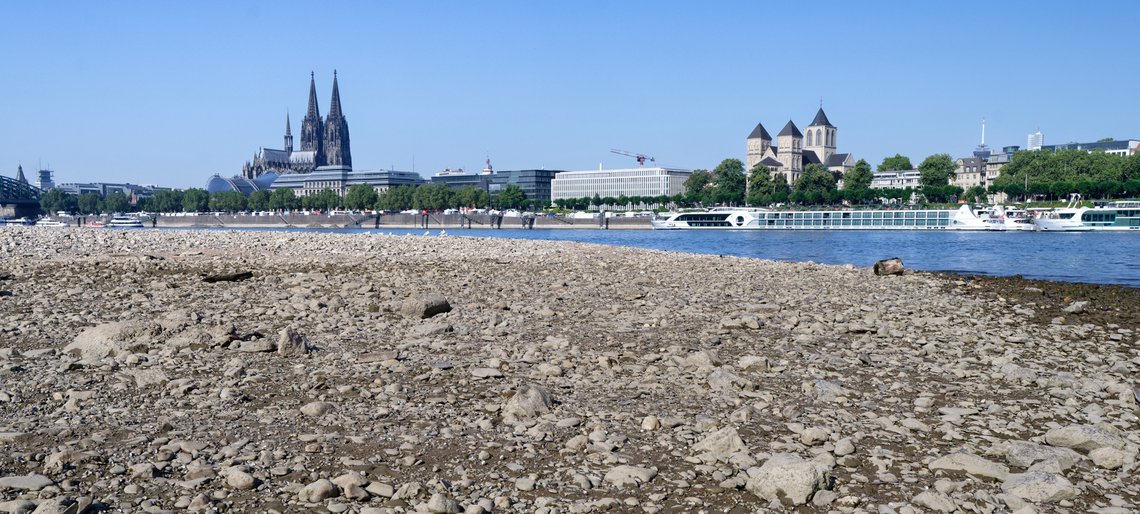 Panorama des Kölner Rheinufers bei Niedrigwasser, mit Dom, Hohenzollernbrücke und Ausflugsschiffen.