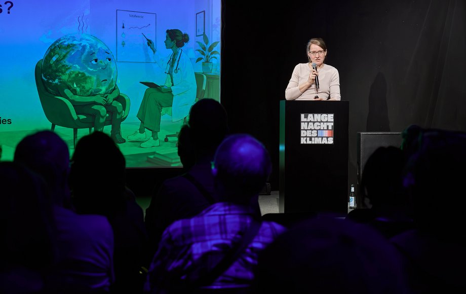Frau auf der Bühne, im Hintergrund ein Cartoon einer kränklichen Erdkugel.