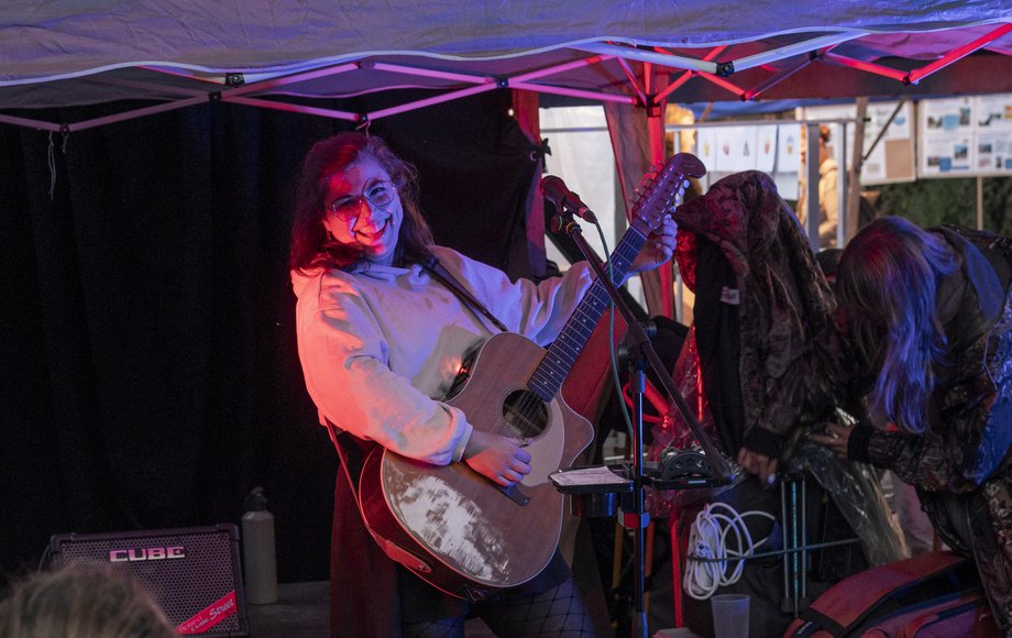 Frau mit Gitarre auf Open-Air-Bühne unter Pavillion in nächtlicher Atmosphäre.