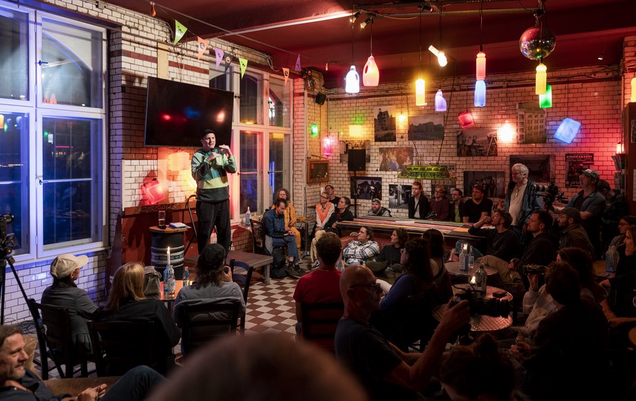 Mann mit Mikrofon steht vor Menschenmenge in einer gefliesten Halle, in der bunte Upcycling-Lampen hängen.