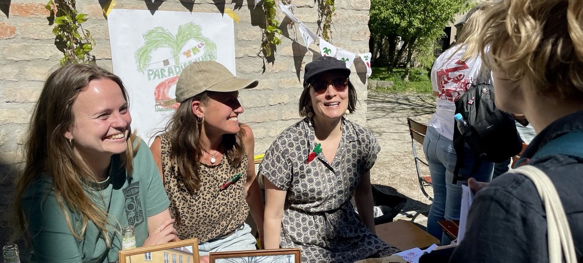 Eine Gruppe von drei lachenden jungen Frauen sitzt an einer Biergarnitur an einem gemauerten Hofeingang und redet mit einer Frau, die von hinten zu sehen ist.