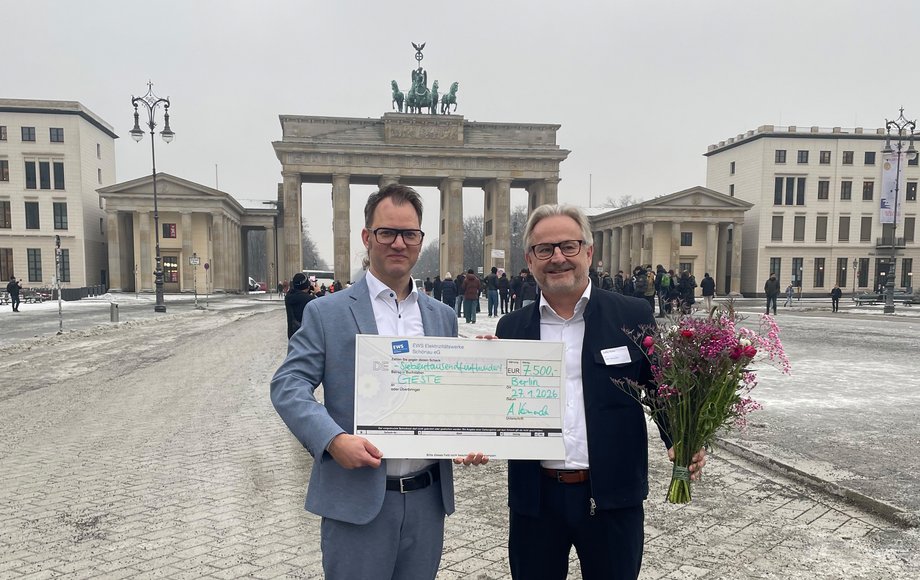 Zwei Menschen stehen vor dem Brandenburger Tor mit einem Spendenscheck und Blumen in der Hand.