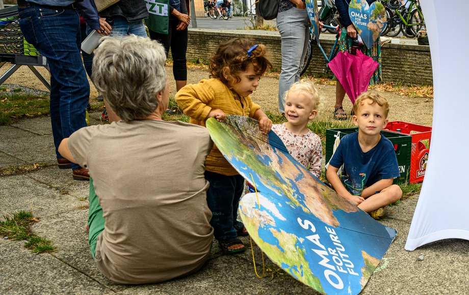 Eine grauhaarige Frau mit dem Schild der Omas For Future hat der Kamera zugewandt; daneben sind drei Kleinkinder