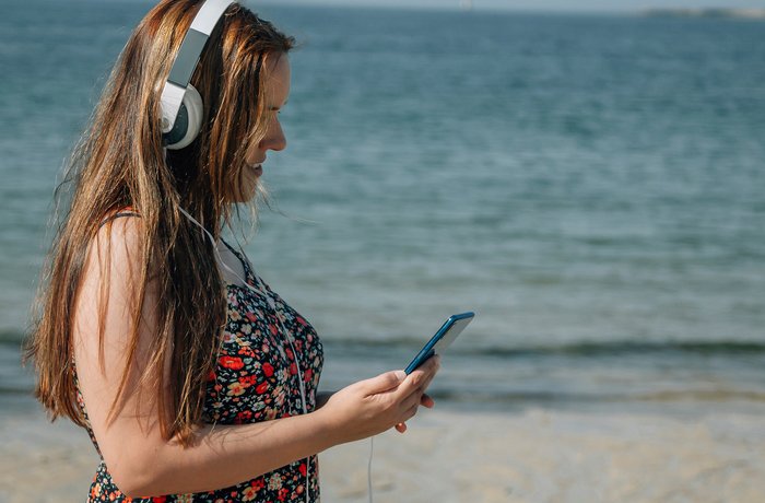 Eine junge Frau im Sommerkleid und mit Kopfhörern auf steht am Strand und schaut auf ihr Mobiltelefon.