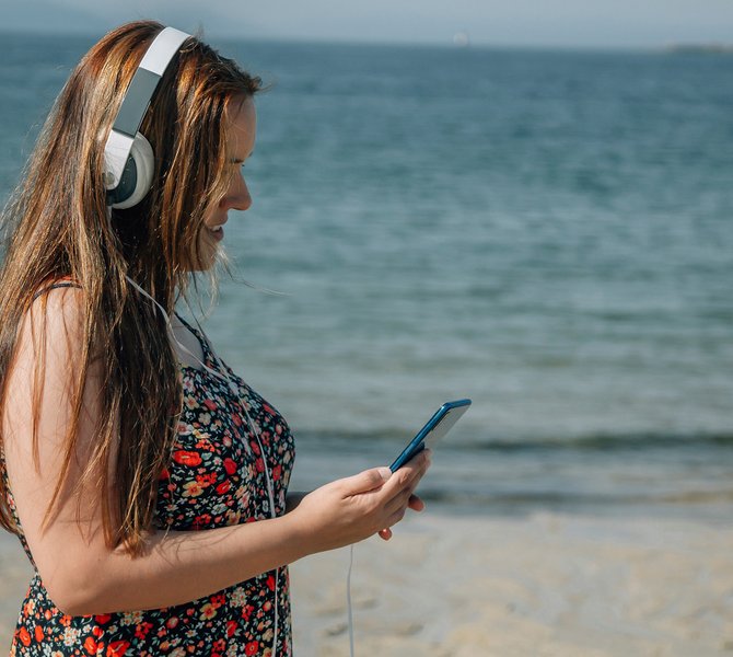Eine junge Frau im Sommerkleid und mit Kopfhörern auf steht am Strand und schaut auf ihr Mobiltelefon.