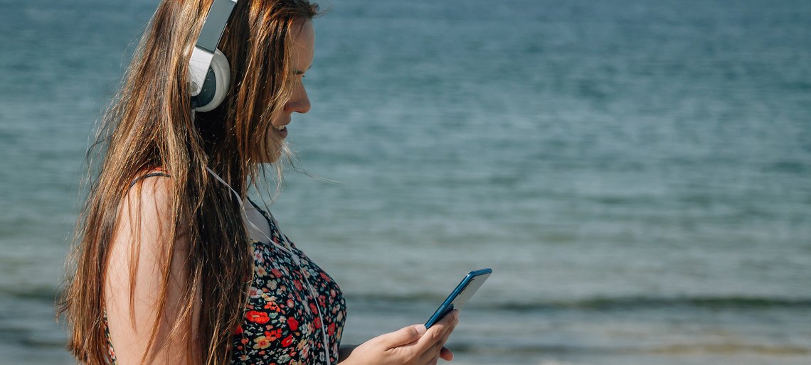 Eine junge Frau im Sommerkleid und mit Kopfhörern auf steht am Strand und schaut auf ihr Mobiltelefon.