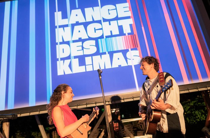Bühne mit zwei Musikern, im Hintergrund auf einer großen Leinwand das Logo