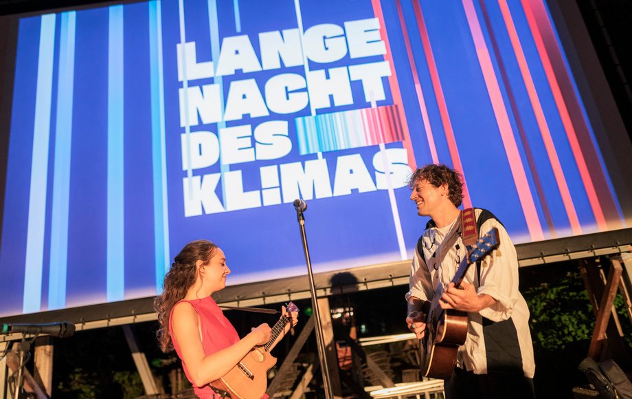 Ein Gitarrist und eine Sängerin auf der Bühne, im Hintergrund ein Warming-Stripes-Backdrop.