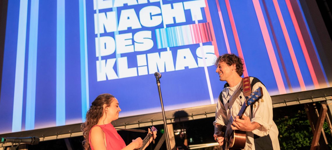 Bühne mit zwei Musikern, im Hintergrund auf einer großen Leinwand das Logo