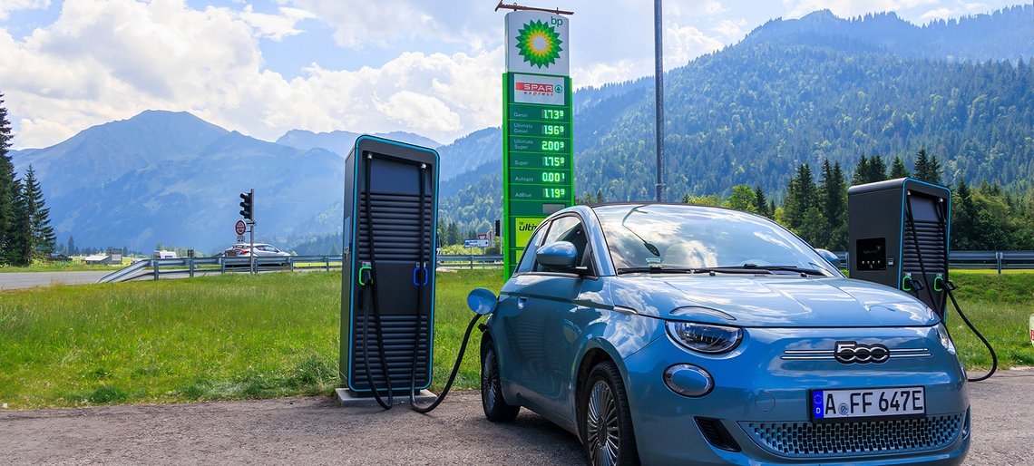 Vor einem Bergpanorama lädt ein E-Auto, im Hintergrund eine Preistafel einer Tankstelle