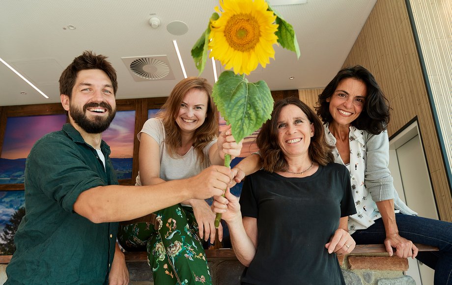 Drei Frauen und ein Mann stehen lachend nebeneinander, gemeinsam halten sie eine große Sonnenblume
