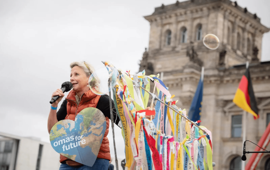 Eine Frau auf der Bühne spricht ins Mikro, hinter ihr flattern viele bunte Bänder