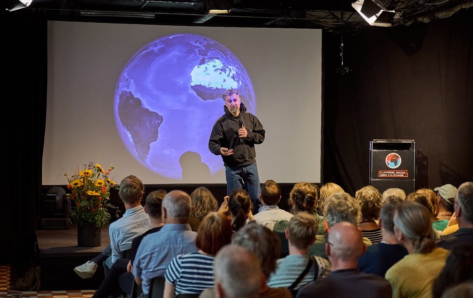 Mann auf der Bühne, im Hintergrund eine projizierte Weltkugel.