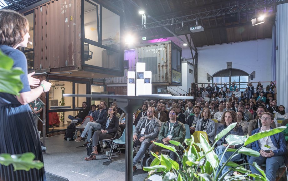 Blick von der Bühne auf eine mit Zuschauern voll besetzte Halle, im linken Bildrand sind Container zu sehen, im Vordergrund Pflanzen