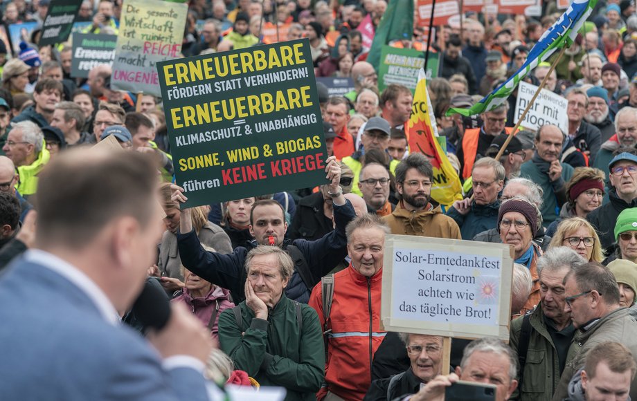 Blick von der Bühne auf verschiedene Demoschilder, Redner von hinten unscharf zu sehen 