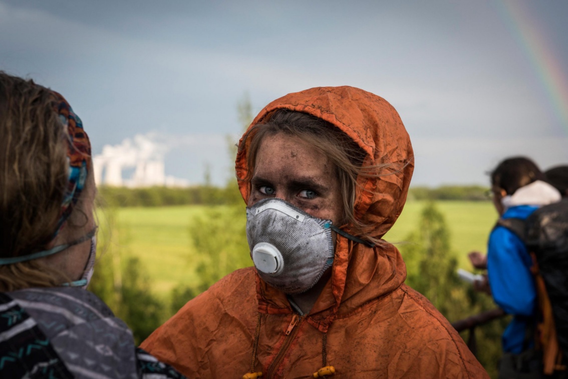 Eine kohlegeschwärzte Aktivistin mit Atemschutzmaske blickt skeptisch-entschlossen in die Kamera.