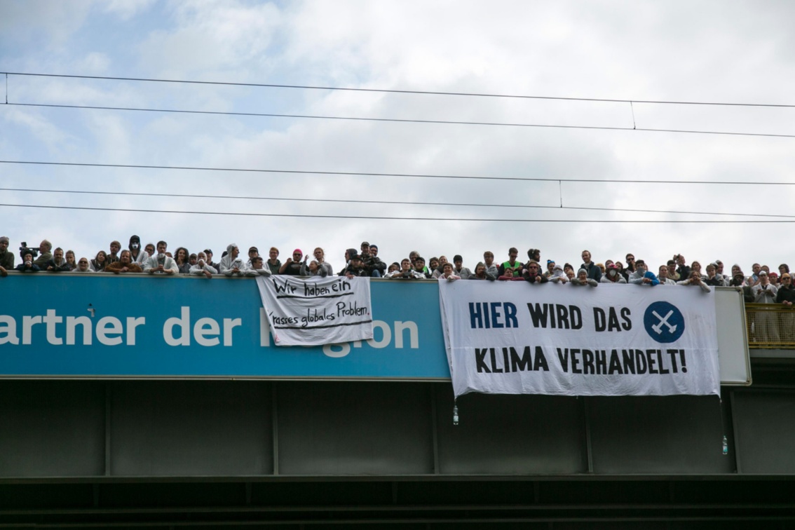 Von einer Brücke, deren Geländer von Demonstranten gesäumt ist, hängen zwei Banner, auf dem größeren steht «Hier wird das Klima verhandelt».