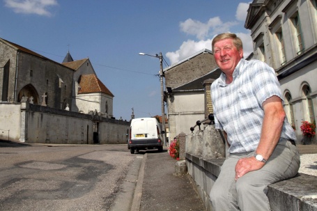 Jean Pierre Remmele, ein fröhlicher älterer Herr mit rosigem Gesicht, sitzt auf einer kleinen Steinmauer an der Dorfstraße.