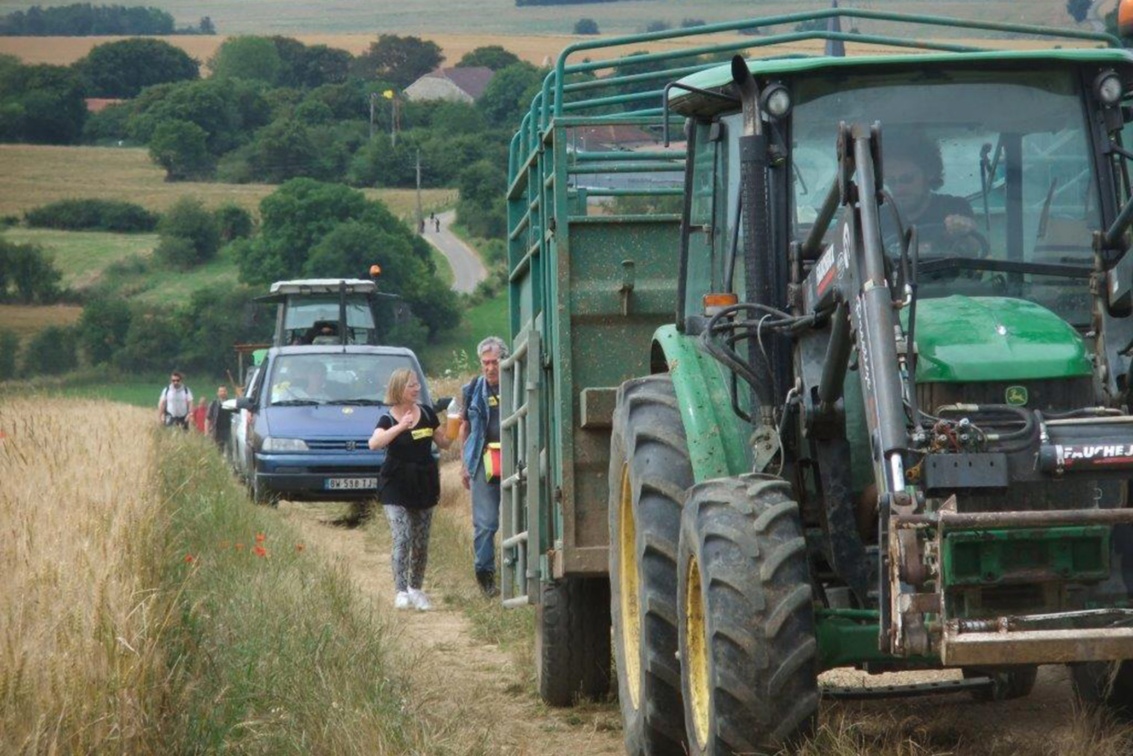 Bewohner begleiten auf einem Feldweg eine Reihe von Fahrzeugen, darunter ein Traktor mit Anhänger.