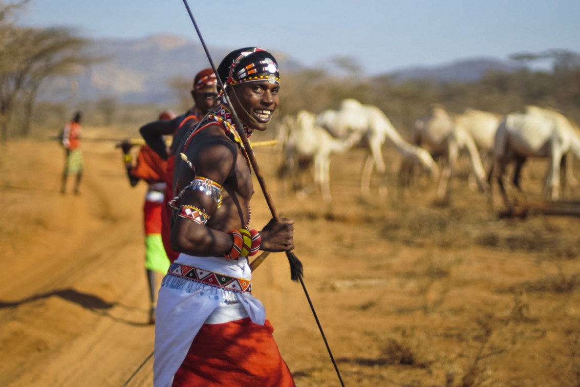 Ein Samburu-Krieger trägt einen Speer, im Hintergrund sind weitere Menschen sowie Kamele zu erkennen.