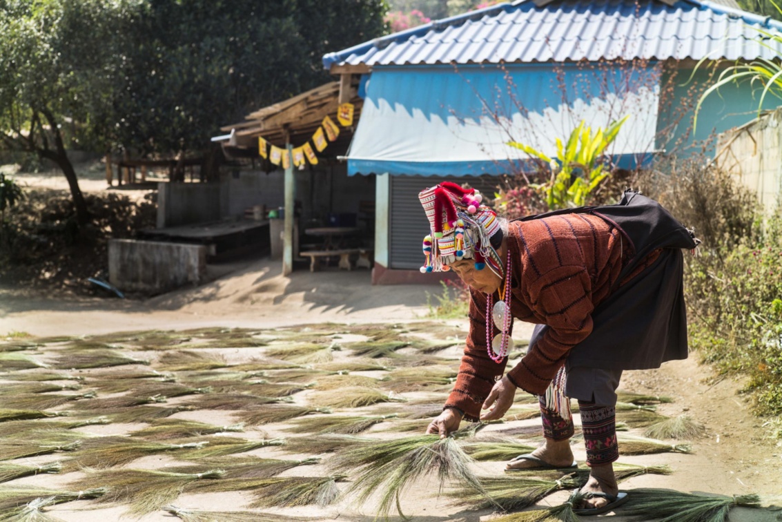 Akha-Frau in traditioneller Tracht legt Grasbesen zum Trocknen auf die Straße