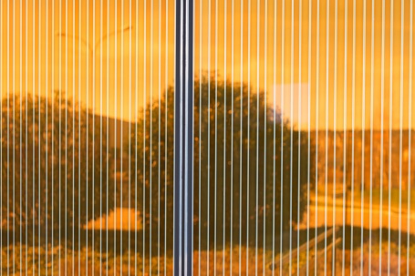 Unscharf spiegelt sich eine baumbestandene Landschaft in einer bronzefarbenen, von hellen Streifen durchzogenen Energieglasfassade. 