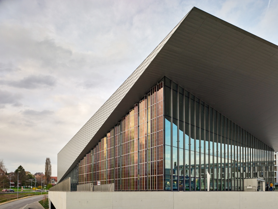 Modernes Gebäude mit kühn auskragendem Dach; eine der Fassadenseiten ist mit Energieglaselementen versehen.