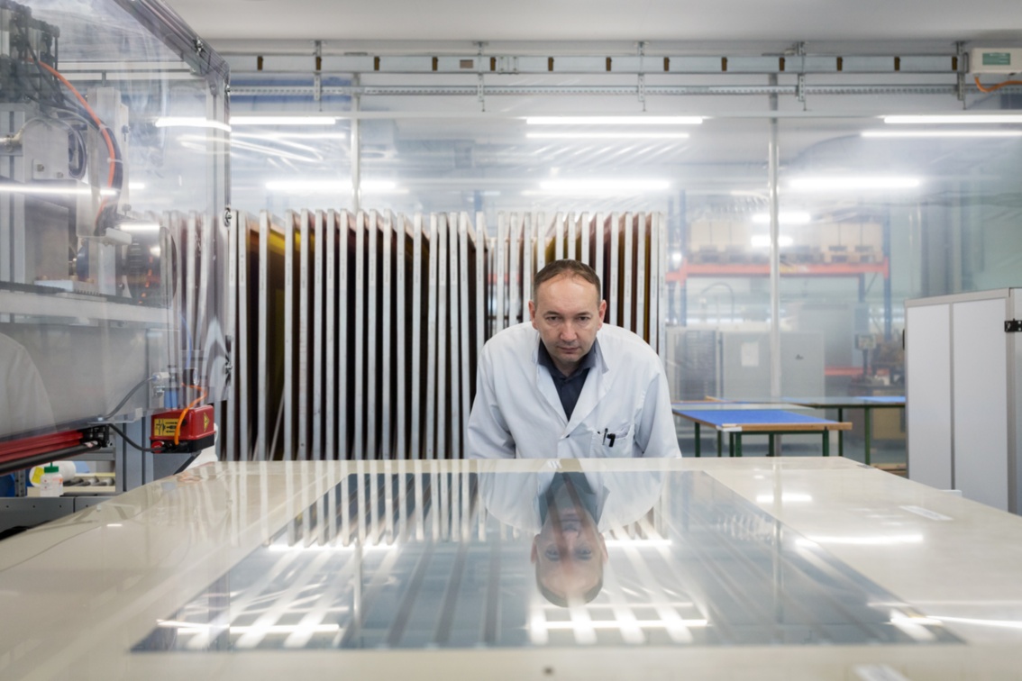 Eine Glasscheibe liegt auf dem Tisch, ein Mann im weißem Laborkittel beugt sich mit prüfendem Blick darüber.