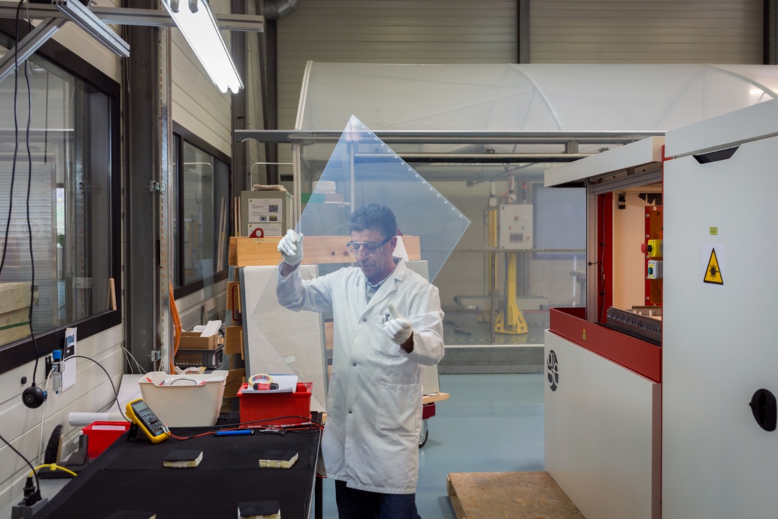 In einer Werkshalle hält ein Mann in weißem Laborkittel eine leicht milchige Glasscheibe mit beiden Armen vor sich.