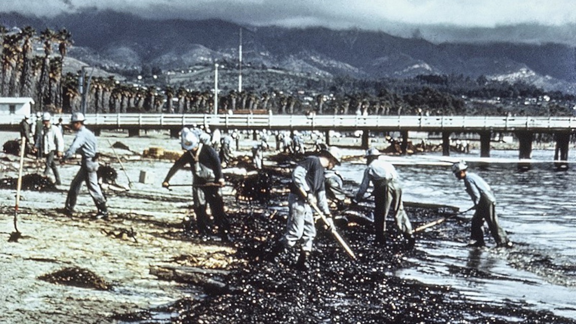 Ein historisches Foto zeigt eine Gruppe Menschen, die mit Schaufeln einen ölverklumpten Strandstreifen säubern.