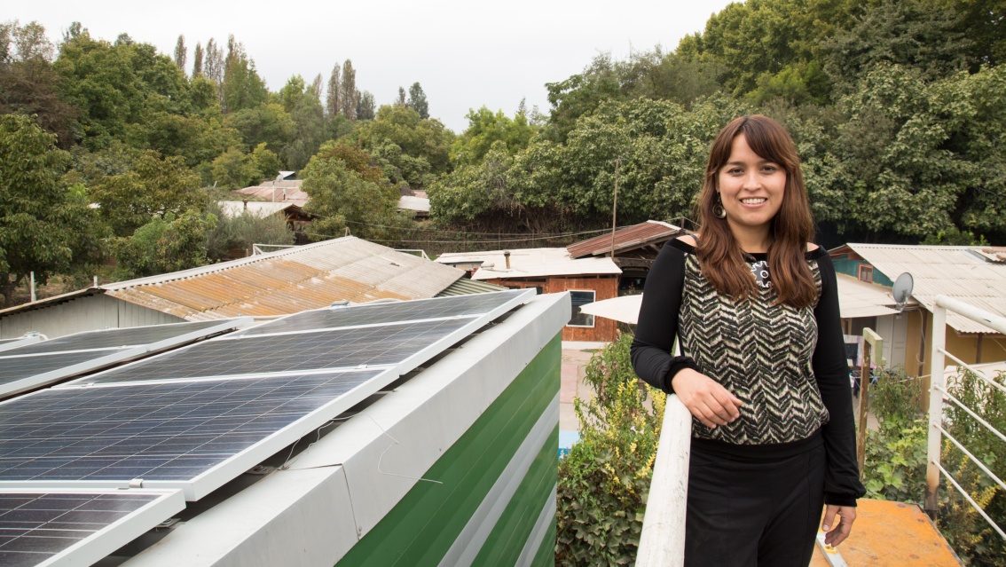 Eine Frau steht lächelnd auf einem Treppengerüst, höhengleich neben ihr ein Dach mit Photovoltaik-Modulen.