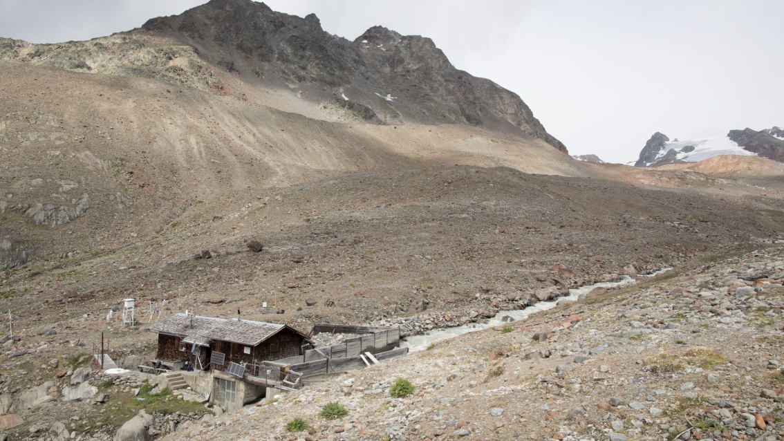 Ein verwittertes Holzhaus mit Steinfundament steht an einem Gletscherbach, der eine von Geröll überzogene Senke durchfließt.