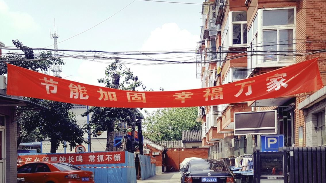 In einer Wohngegend mit geparkten Autos überspannt ein rotes Banner mit chinesischen Schriftzeichen einen Zufahrtsweg.
