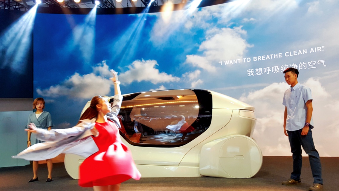 Automobilmesse: Vor der aerodynamischen Karosserie einer automobilen Konzeptstudie tanzt eine Frau in rotem Kleid.