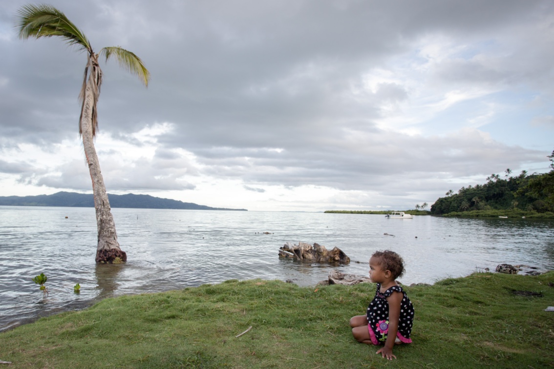 Ein Kind sitzt im Gras am am Meer, im Wasser steht eine Palme.