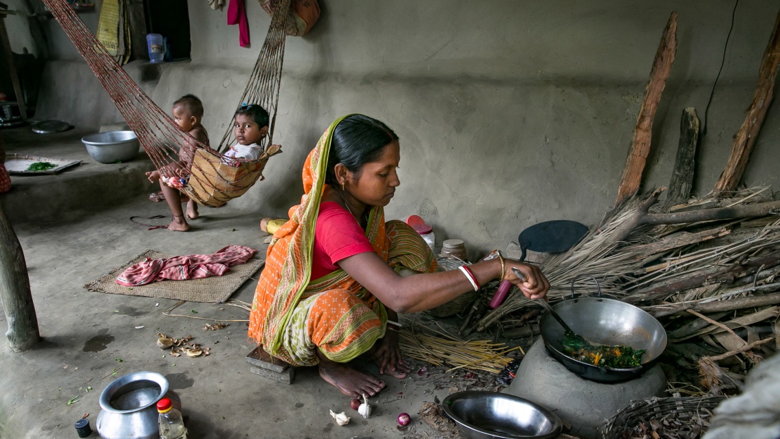 Einer Mutter sitzt auf dem Boden und kocht, im Hintergrund zwei kleine Kinder in einer Hängematte.