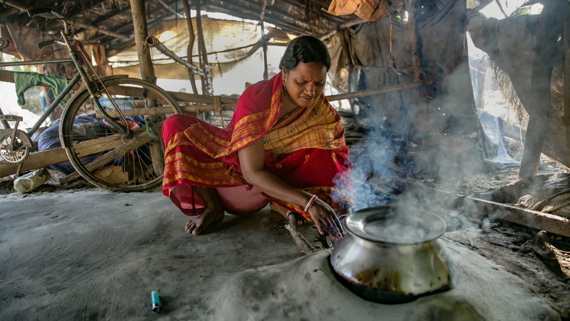 Eine hübsche Frau in einem leuchtend roten Sari kocht vor einer qualmenden Feuerstelle in einer ärmlichen Behausung