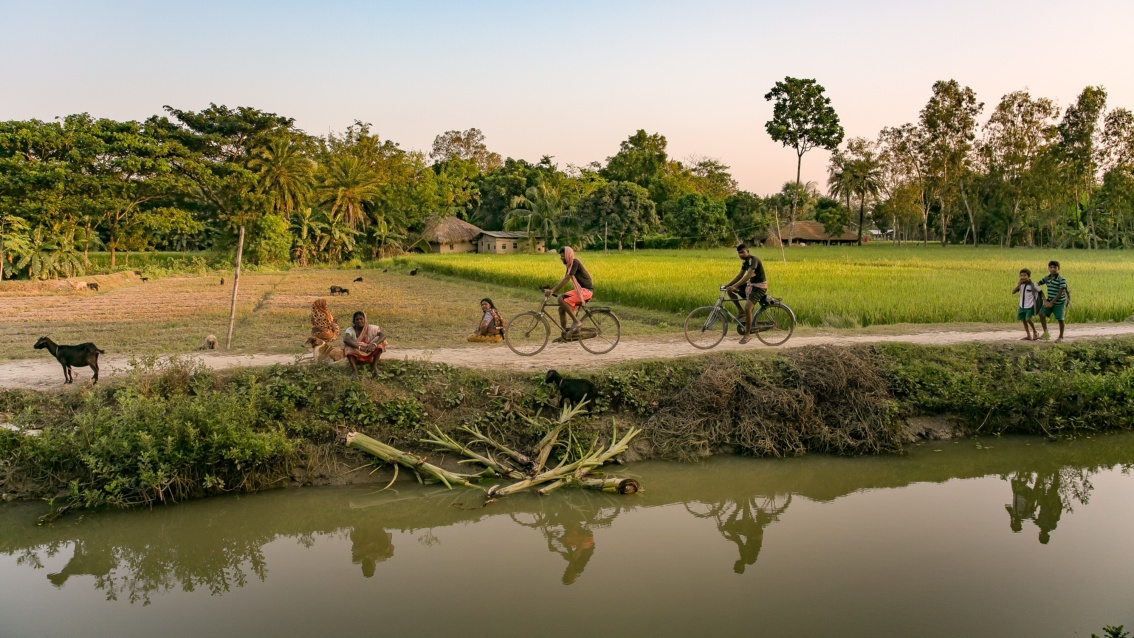 An einem Fluß, im Hintergrund ein Reisfeld und ein Weg, auf dem Fahrräder unterwegs sind.