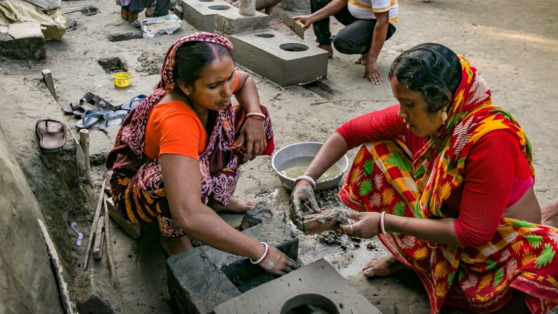 Zwei Frauen in leuchtenden Saris formen Lehm für die vor ihnen stehende Lehmherd-Grundform