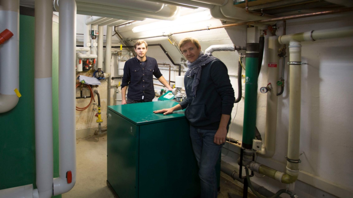 Zwei Mitarbeiter zeigen eine KWK-Anlage in einem Kellerraum.