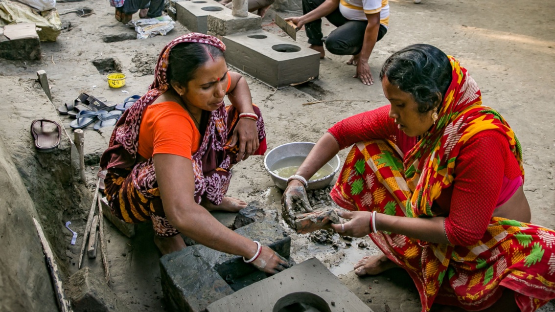 Zwei indische Frauen in roten Saris bauen gemeinsam einen Lehmherd