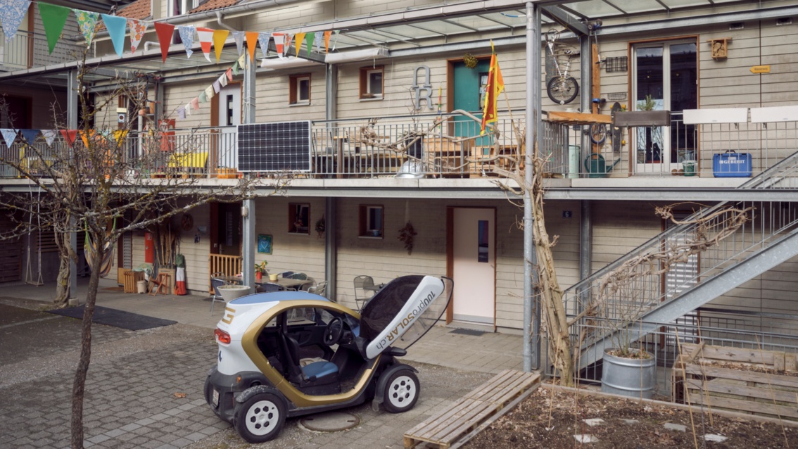 Eine Häuserzeile mit offenen Balkonen und bunten Fahnen, davor steht ein Elektroauto.