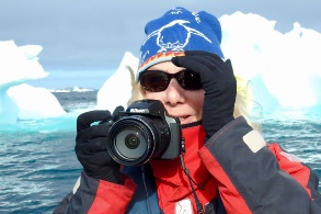 Daphne Bramham mit Mütze, Sonnenbrille und Kamera hält ihre behandschuhten Hände schützend an die Schläfen, im Hintergrund Eisberge.