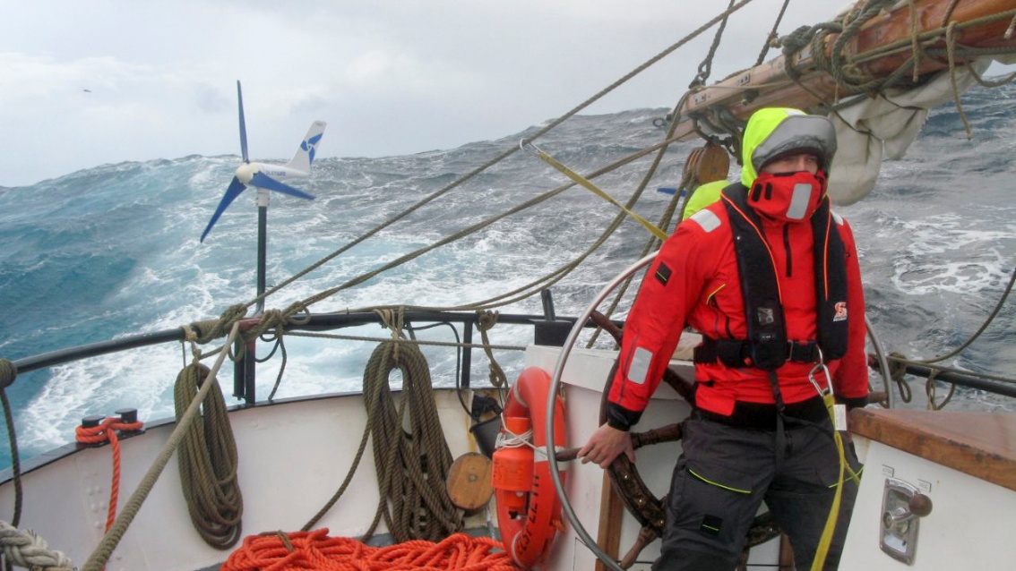 Mit Segelkleidung gegen Wind und Wasser geschützt, hält ein Besatzungsmitglied das Steuerrad; dahinter meterhoch aufschäumende Wellen.