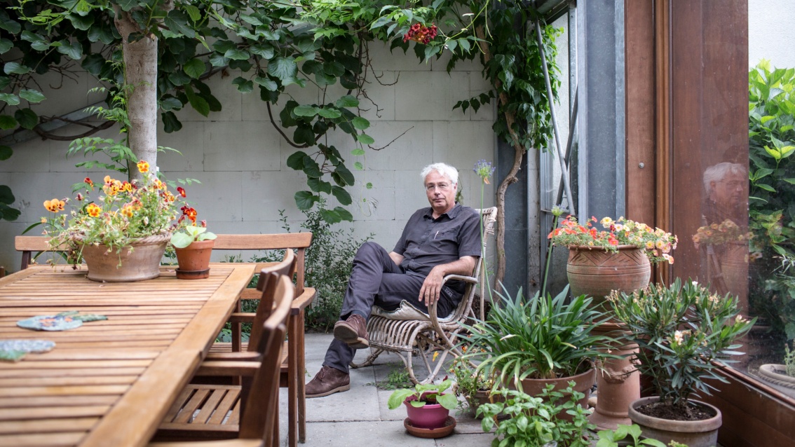 Entspannt sitzt ein älterer Herr mit übereinandergeschlagenen Beinen in einem Rattansessel neben einem Holztisch. Vor der unverputzten Wand hinter ihm wächst eine baumgroße Pflanze.