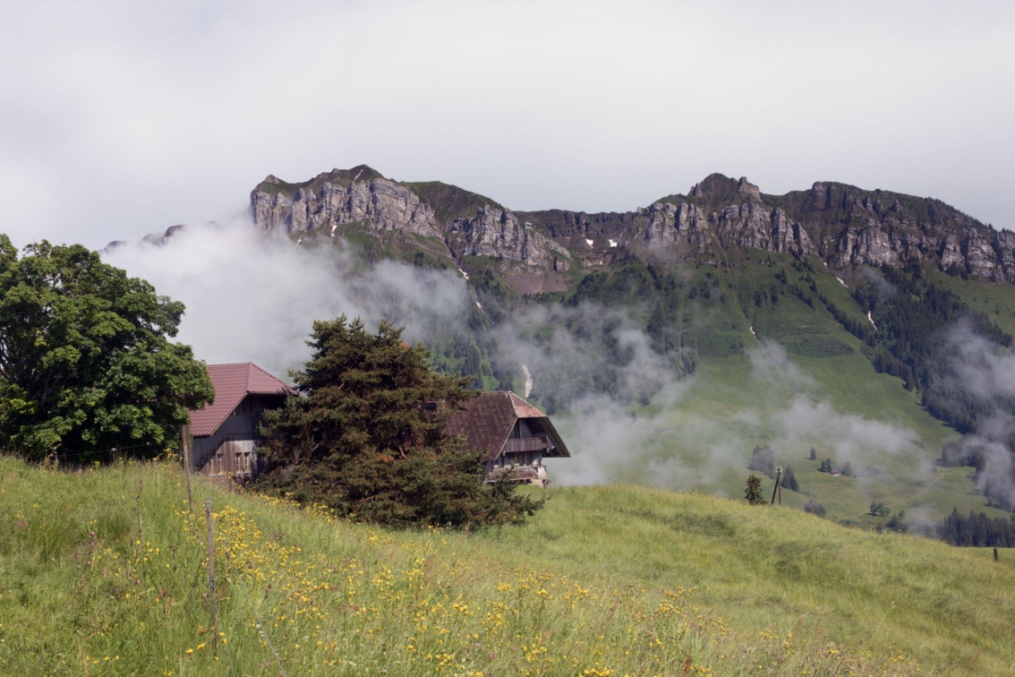  Auf einer Bergwiese stehen eine Alp aus zwei Höfen mit Walmdach,  dahinter geben wegziehende Wolken den Blick auf eine Bergkette frei.