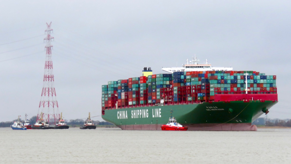 Ein riesiges , bis oben beladenes Containerschiff, umgeben von diversen Schleppern, im Hintergrund ein rot-weißer Strommast
